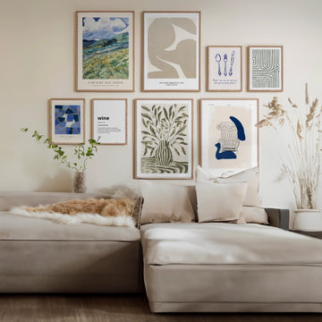 Een moderne woonkamer is voorzien van een lichtgrijze bank met witte en beige kussens, een potplant en een decoratief arrangement van gedroogde planten. De muur toont een galerij met ingelijste kunstafdrukken van gerenommeerde kunstenaars, waaronder landschappen, abstracte ontwerpen en op tekst gebaseerde kunstwerken uit Mixtiles Set No. 8 van Collage Depot B.V.