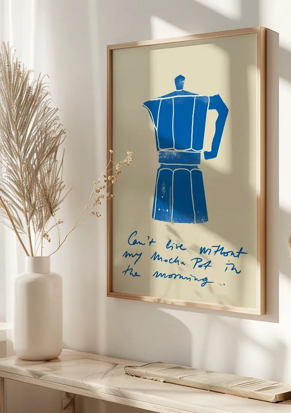 Aan de muur hangt een ingelijste kunstprint met een blauwe Moka Pot-illustratie, vergezeld van een handgeschreven tekst met de tekst: "Kan niet leven zonder mijn Mokkapot in de ochtend..." Het Can't Live Without My Moka Pot-schilderij van CollageDepot staat naast een witte vaas met gedroogde planten.