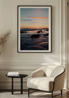 Een ingelijste foto van een strand zonsondergang Langs Het Water Schilderij van CollageDepot hangt aan een lichtgekleurde muur. Onder het schilderij staat een moderne witte fauteuil en een klein zwart rond bijzettafeltje met een stapel boeken. De kamer is voorzien van elegant lijstwerk en een gedroogde plantenopstelling aan de linkerkant, waardoor een perfecte wanddecoratie ontstaat.,Zwart