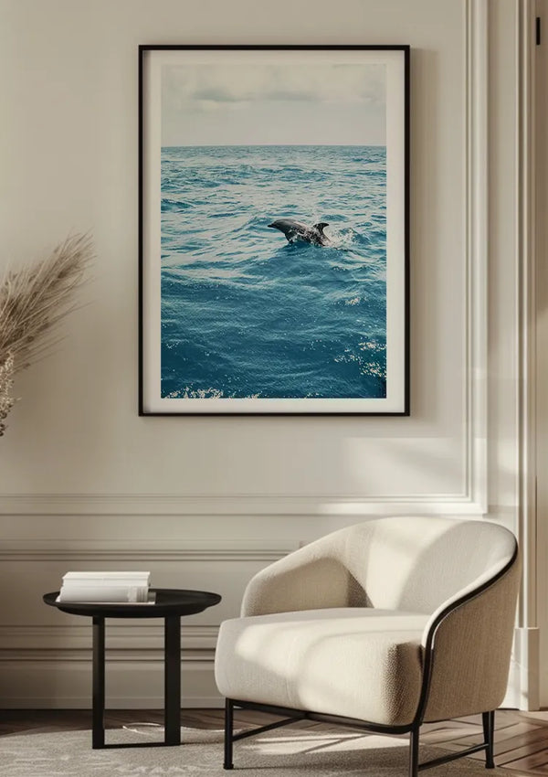 Aan een beige muur hangt een ingelijste foto van een walvis die in de oceaan opduikt als CollageDepot Springende Dolfijn In De Natuur Schilderij. Onder de foto staat een witte fauteuil en een ronde zwarte bijzettafel met een boek en een vaas. Aan de linkerkant is gedroogd pampagras decoratief geplaatst, wat het serene tafereel compleet maakt.,Zwart
