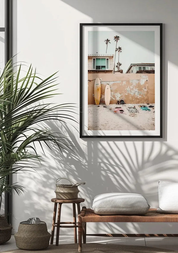 Een ingelijst Surfbord Op Het Strand Schilderij van CollageDepot hangt aan een witte muur. Daaronder staat een houten bank met daaronder rieten manden en links een potplant. De foto toont surfplanken en palmbomen bij een zandstrand met strandlakens, waardoor een perfecte wanddecoratie ontstaat.,Zwart