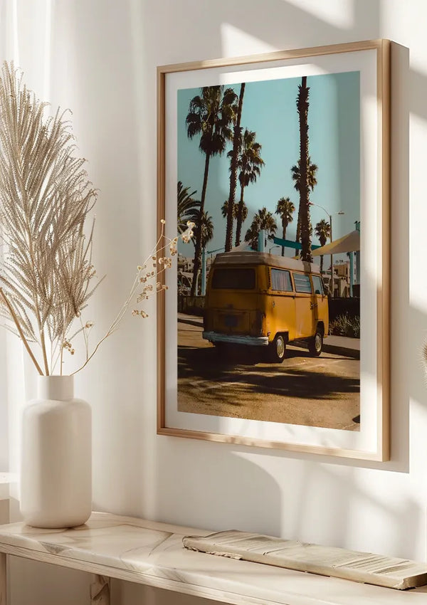 Ingelijste foto van een CollageDepot Vintage Busje Omringd Door Palmbomen, hangend aan de muur met een magnetisch ophangsysteem. Vlakbij staat een witte vaas met daarin gedroogde planten, op een witte plank geplaatst. Natuurlijk licht filtert naar binnen en werpt schaduwen op de muur, waardoor deze unieke wanddecoratie nog beter tot zijn recht komt.,Lichtbruin