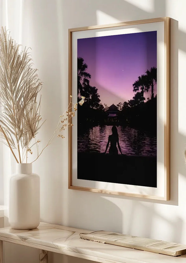 Aan een witte muur hangt een ingelijste foto van een tropische zonsondergang, met een silhouet van een persoon bij een zwembad met palmbomen en een paviljoen op de achtergrond. Dit Silhouet Aan Het Zwembad Schilderij van CollageDepot, gevat in een houten frame, staat naast een decoratieve vaas met gedroogde planten en maakt gebruik van een ingenieus magnetisch ophangsysteem.,Lichtbruin