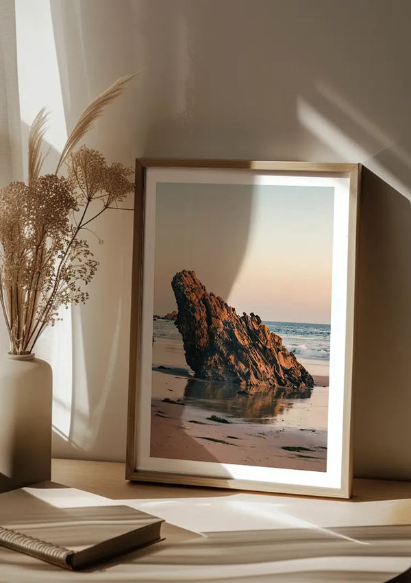 Een ingelijste Uitstekende Rots Schilderij van CollageDepot van een rotsformatie aan de kust op een zandstrand is op een houten oppervlak geplaatst. Ernaast staat een vaas met gedroogde planten en een opengeslagen boek. Zonlicht filtert door transparante gordijnen en werpt zachte schaduwen, wat de serene sfeer van dit wanddecoratiearrangement versterkt.,Lichtbruin