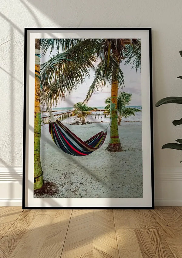 Op een houten vloer rust een ingelijste foto met een hangmat, vastgebonden tussen twee palmbomen op een zandstrand. Op de achtergrond steekt een houten pier uit in de oceaan, en verspreid over het strand staan nog meer palmbomen. Dit stukje wanddecoratie, Hangen Tussen De Palmen Schilderij van CollageDepot, kan eenvoudig worden tentoongesteld met een magnetisch ophangsysteem.,Zwart