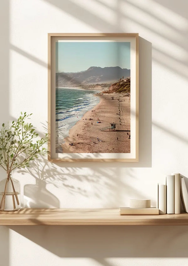 Een ingelijste foto van een strand met mensen op het zand en de golven van de oceaan, afgezet tegen de bergen, wordt weergegeven als een prachtig CollageDepot Geweldig Uitzicht Schilderij op een houten plank. Op de plank staat ook een kleine plant en een paar boeken. Zonlicht zorgt voor schaduwen op de muur achter de plank.,Lichtbruin