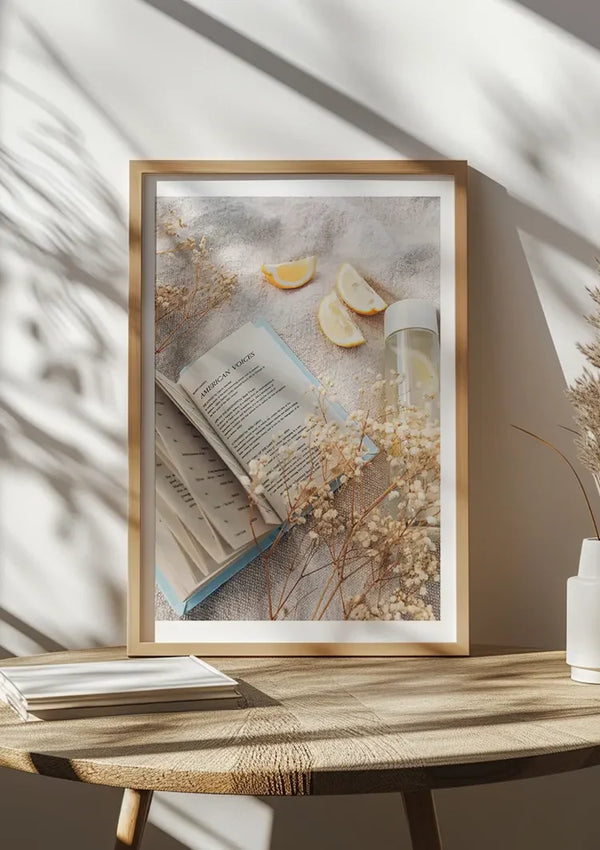 Ingelijste foto op een houten tafel met een open boek, partjes citroen, een spuitfles en gedroogde bloemenelementen. Zonlicht werpt schaduwen op het tafereel en voegt een luchtige sfeer toe. Aan de linkerkant van de tafel is gedeeltelijk een stapel boeken zichtbaar. Dit serene "Ontspannen met een boekschilderij" van CollageDepot legt een moment in de tijd vast.