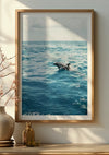 Een ingelijste foto van een dolfijn die in de oceaan zwemt, wordt met behulp van een magnetisch ophangsysteem aan een beige geschilderde muur gehangen. Het serene tafereel laat zien dat zonlicht schaduwen werpt. Een vaas met een minimalistisch bloemstuk en een klein glazen flesje staan op een plank onder dit elegante Springende Dolfijn In De Natuur Schilderij van CollageDepot.,Lichtbruin