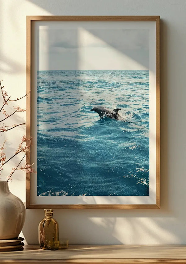 Een ingelijste foto van een dolfijn die in de oceaan zwemt, wordt met behulp van een magnetisch ophangsysteem aan een beige geschilderde muur gehangen. Het serene tafereel laat zien dat zonlicht schaduwen werpt. Een vaas met een minimalistisch bloemstuk en een klein glazen flesje staan op een plank onder dit elegante Springende Dolfijn In De Natuur Schilderij van CollageDepot.,Lichtbruin