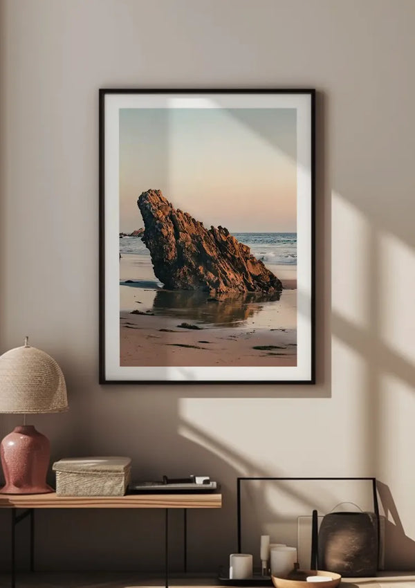 Aan een beige muur hangt een ingelijste foto van een grote rotsformatie op een strand, vastgehouden door een magnetisch ophangsysteem. Het strandtafereel toont de rots die bij zonsondergang uit het zand bij de oceaan steekt. Onder deze prachtige wanddecoratie, *Uitstekende Rots Schilderij* van *CollageDepot*, staat een tafel met een lamp, een doos en enkele decoratieve items.,Zwart