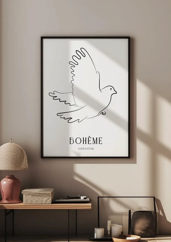 Aan een beige muur hangt een ingelijste minimalistische lijntekening van een vogel met daaronder de tekst "BOHÈME collection". Onder het CollageDepot Bohème Collection Schilderij staat een tafel met een roze lamp, een doos en andere kleine spullen. Zonlicht werpt schaduwen op de muur.,Zwart