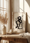 Een ingelijst abstract zwart-wit Grafisch figuur schilderij van CollageDepot staat op een houten tafel bij een raam. Op de tafel staat ook een keramische vaas met gedroogd blad, een kleine kruik, een stapel boeken en een gedrapeerd kleed. Zonlicht stroomt door de vitrages en werpt schaduwen op de elegante wanddecoratie.