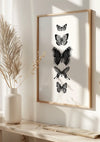 Een ingelijst kunstwerk van vijf monochrome vlinders die aan een witte muur hangen. Bij de afbeelding hoort een witte vaas met gedroogde planten, op een lichtgekleurde plank eronder geplaatst. Fel zonlicht werpt schaduwen op de muur en plank, en het magnetische ophangsysteem zorgt voor een moeiteloze presentatie als elegante wanddecoratie. Het product heet Vijf Verschillende Vlinders Op Een Rij Schilderij van CollageDepot.,Lichtbruin