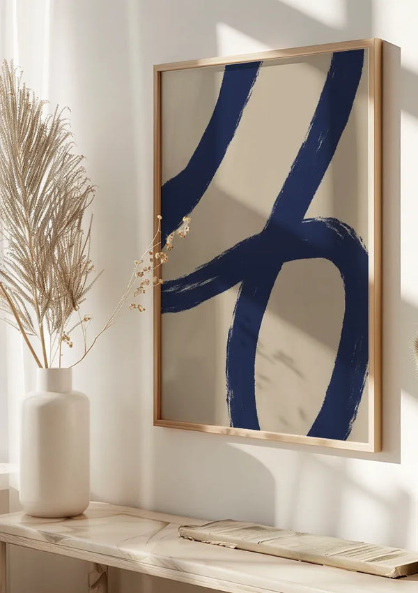 Een minimalistisch Blue Octagonal Loop-schilderij van CollageDepot met opvallende blauwe penseelstreken op een beige achtergrond, ingelijst in licht hout. Het wordt aan een lichtgekleurde muur gehangen boven een marmeren oppervlak met een witte vaas met gedroogde siergrassen. Licht filtert er van links doorheen.,Lichtbruin