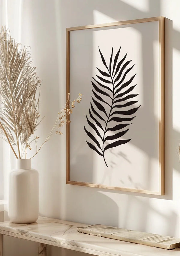 Een minimalistische kamer is voorzien van een ingelijste zwart-witte botanische print van een enkel blad op een witte muur als stijlvolle wanddecoratie. Een witte vaas met gedroogd pampagras en fijne takjes staat op een houten oppervlak onder het schilderij Zwarte palm van CollageDepot. Natuurlijk licht werpt zachte schaduwen.