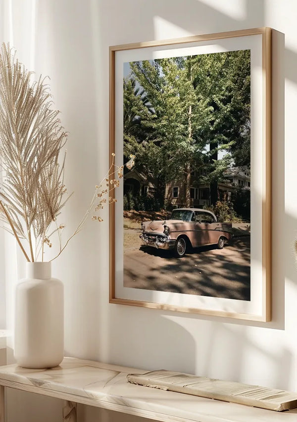 Aan een witte muur hangt een ingelijste foto met een afbeelding van een vintage roze auto die geparkeerd staat voor een huis met bomen. Dit Roze Vintage Auto Schilderij van CollageDepot wordt tentoongesteld in een houten frame boven een lichtgekleurde plank versierd met gedroogde bloemstukken in witte vazen.,Lichtbruin