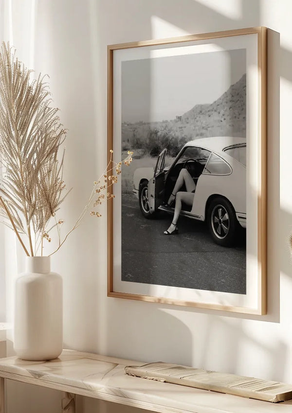 Aan de muur hangt een ingelijste zwart-witfoto, een prachtig Porsche With Love Schilderij van CollageDepot, met een afbeelding van een persoon die op de bestuurdersstoel van een oldtimer zit met de deur open. Onder de foto, weergegeven door middel van een magnetisch ophangsysteem, staat een houten plank met een vaas met decoratieve gedroogde planten.,Lichtbruin