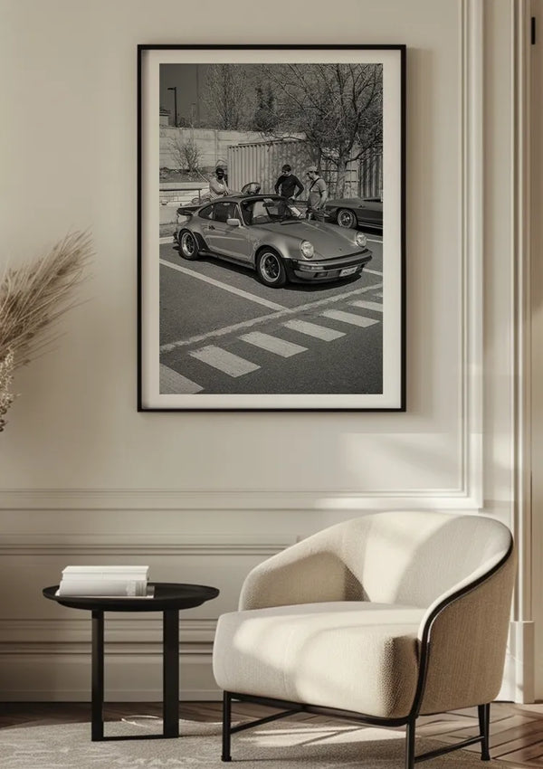 Een ingelijste zwart-witfoto van een Klassieke Porsche 911 Turbo Schilderij van CollageDepot is gemonteerd op een witte muur boven een beige fauteuil en een zwarte ronde bijzettafel. De kamer heeft een minimalistische inrichting met moderne esthetische en natuurlijke verlichting, waardoor een elegant middelpunt van de wanddecoratie ontstaat.,Zwart