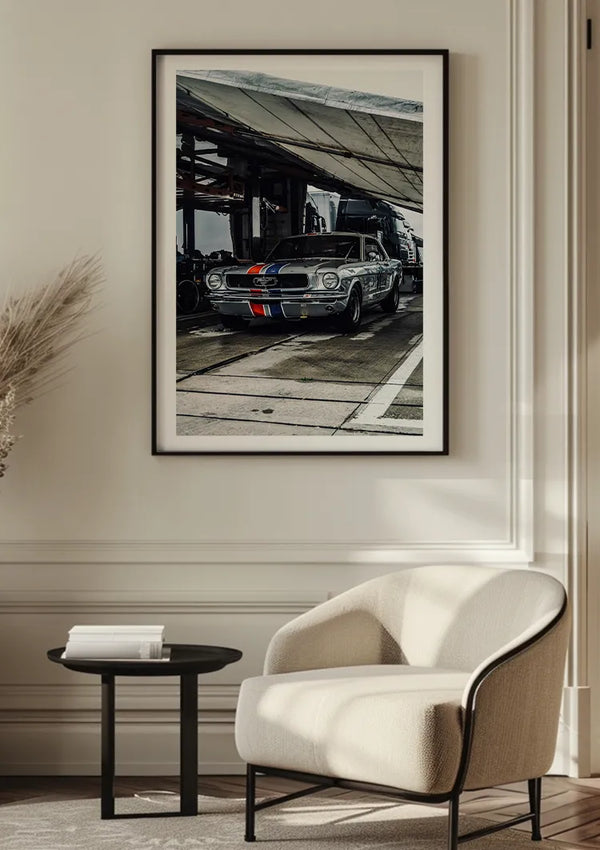Een ingelijste foto van een vintage Ford Mustang met een blauwe en rode streep op een beige muur. Onder het frame bevindt zich een moderne witte fauteuil en een ronde zwarte bijzettafel. Dit stijlvolle Zilveren Ford Mustang Schilderij van CollageDepot voegt een moeiteloos chique tintje toe aan uw ruimte.,Zwart