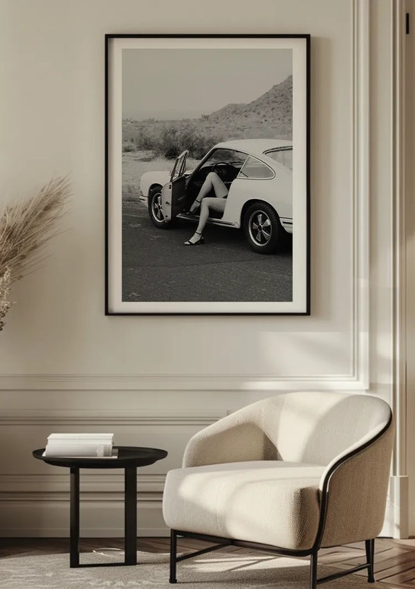 Een ingelijste zwart-wit foto, het Porsche With Love Schilderij van CollageDepot, een prachtige wanddecoratie, is gemonteerd op een beige muur. Op de foto is een persoon te zien die in een geparkeerde oldtimer zit met de deur open. Voor de foto staat een moderne witte fauteuil en een klein zwart bijzettafeltje met boeken.,Zwart