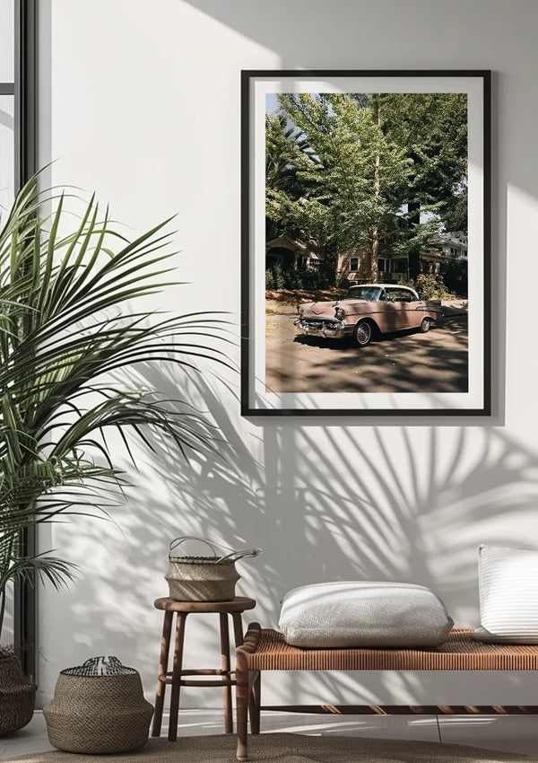 Een ingelijste foto van een Roze Vintage Auto Schilderij van CollageDepot, geparkeerd in een met bomen omzoomde straat, wordt als elegante wanddecoratie op een witte muur getoond. Onder de foto staat een houten bank met kussens en een mand, terwijl zonlicht schaduwen werpt van een grote potplant.,Zwart
