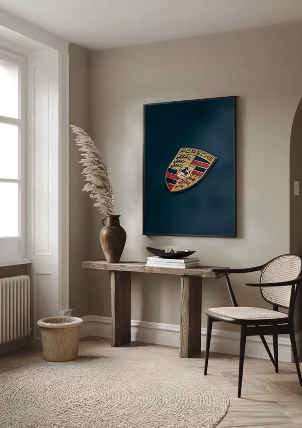 Een moderne, minimalistische kamer met een houten consoletafel tegen een beige muur. Boven de tafel hangt een ingelijst Porsche Logo Schilderij van CollageDepot. Op de tafel staat een grote vaas met gedroogd pampagras, gestapelde boeken en een zwarte kom. Naast de tafel staan een rieten mand en een stoel, die de smaakvolle wanddecoratie versterken.,Zwart