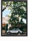 Een zilveren vintage Zilveren Porsche 911 Oldtimer Schilderij van CollageDepot staat geparkeerd in een straat vol weelderig groen en hoge bomen. Op de achtergrond is een huis met een veranda en pannendak zichtbaar, gedeeltelijk in de schaduw van overhangende takken. De scène is helder en zonovergoten, een perfecte setting voor een schilderij of wanddecoratie.,Zwart-Zonder,Lichtbruin-Zonder,showOne,Zonder