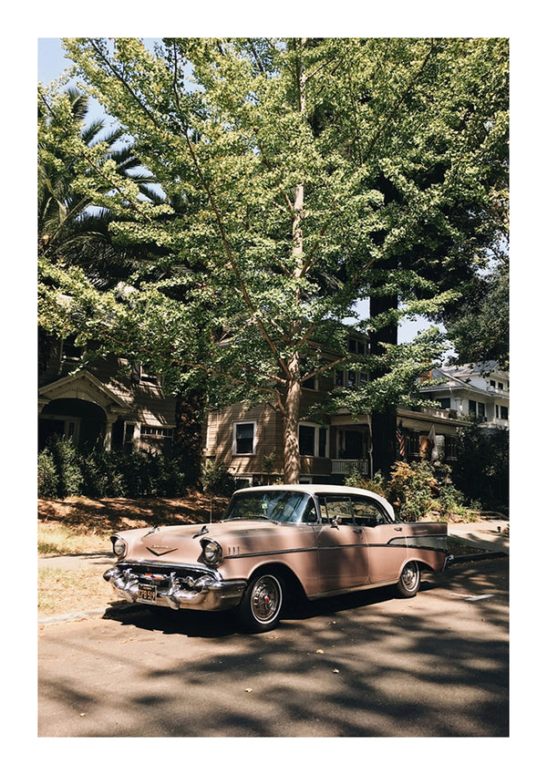 Aan de zijkant van een woonstraat hangt een lichtbruin Roze Vintage Auto Schilderij van CollageDepot. Op de achtergrond zijn huizen te zien die gedeeltelijk aan het zicht worden onttrokken door grote groene bomen, wat duidt op een buitenwijk. Het weer ziet er zonnig uit met een heldere hemel.-