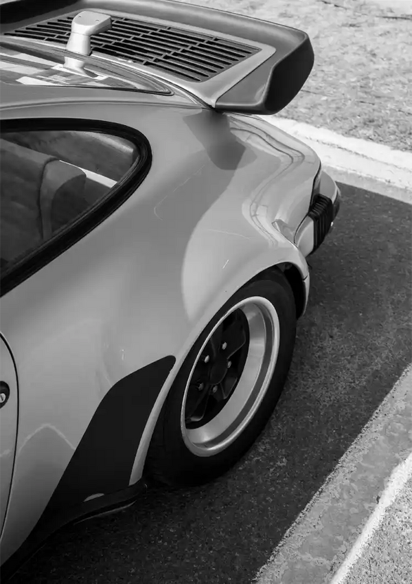 Een zwart-witfoto van de achterkant van een sportwagen, waarbij de nadruk ligt op het ronde ontwerp, de grote wielen en de opvallende achterspoiler, geparkeerd langs een weg bij de aaa 076 - auto's van CollageDepot.-