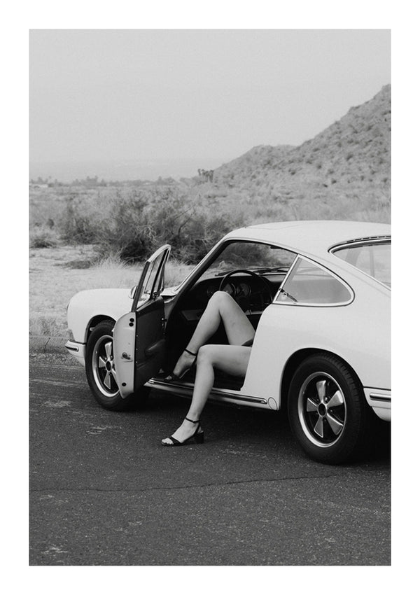 Zwart-witfoto van een persoon die in een CollageDepot aaa 098 zit - auto's met de deur open, alleen de benen zichtbaar. De auto staat langs de kant van de weg geparkeerd met een dor landschap op de achtergrond.-