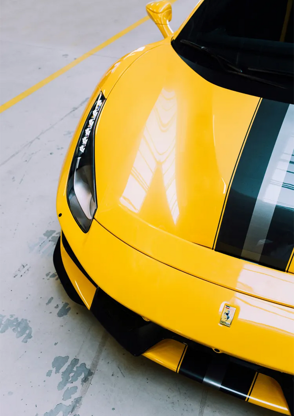 Een bovenaanzicht van de voorkant van een gele sportwagen met zwarte racestrepen, geparkeerd in een garage met betonnen vloer. De afbeelding toont de linkerkoplamp, een deel van de motorkap en het logo van de iconische autofabrikant. Deze scène wordt perfect weergegeven in "aaa 119 - auto's" van CollageDepot.-