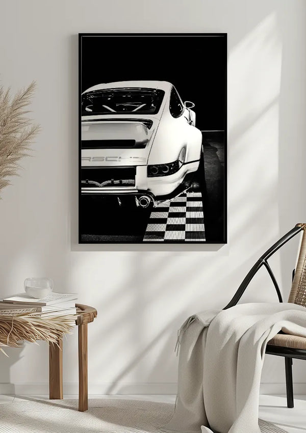 Een minimalistisch interieur met een Zwart-Witte Porsche Schilderij van CollageDepot aan de muur. Vlakbij staat een rieten stoel met een gedrapeerde deken en een tafeltje, versierd met witte boeken en decoratieve voorwerpen. Neutrale tinten en natuurlijk licht domineren de ruimte.,Zwart