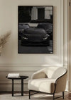 Een Zwart op Zwart Lamborghini Schilderij van CollageDepot hangt aan een beige muur boven een moderne, crèmekleurige fauteuil en een kleine, ronde zwarte tafel. De scène speelt zich af in een minimalistische woonkamer met een elegante inrichting, waaronder een vaas met gedroogd blad.,Zwart