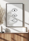 Een ingelijste poster met drie gestileerde oogillustraties van CollageDepot's product 002 - bestsellers, hangend aan een muur boven een houten plank met decorartikelen, en werpt een zachte schaduw in een zonovergoten kamer.,Zwart