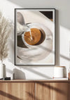 Een ingelijst Cappuccino-schilderij van CollageDepot, hangend aan een witte muur boven een houten kast. De afbeelding toont een illustratie van melk die in een kopje koffie wordt gegoten. In de kast staan decoratieve spullen zoals vazen en een plantje. Zonlicht werpt schaduwen over het tafereel en versterkt de elegante wanddecoratie.,Zwart