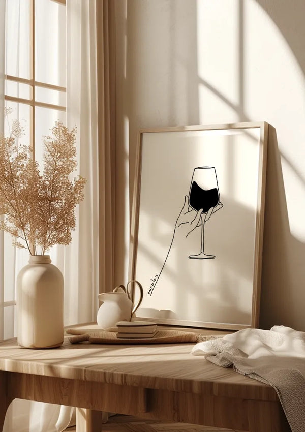 Een ingelijste lijntekening van een hand die een wijnglas vasthoudt, rust op een houten tafel versierd met **CollageDepot's Abstract Wine Glass Schilderij**. Op de tafel staat ook een vaasje met gedroogde bloemen, een theepot, twee boeken en een opgevouwen wit kleed. Zonlicht stroomt door een nabijgelegen raam en werpt schaduwen op deze elegante wanddecoratie-opstelling.,Lichtbruin