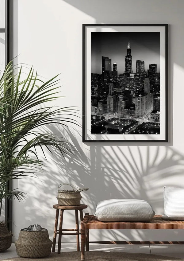 Een ingelijste zwart-witfoto van een stadsgezicht, de Skyline At Night Schilderij van CollageDepot, wordt weergegeven als een prachtig stukje wanddecoratie op een witte muur. Onder de foto staat een houten bank met kussens. Links werpt een grote bladplant schaduwen op de muur en de vloer.