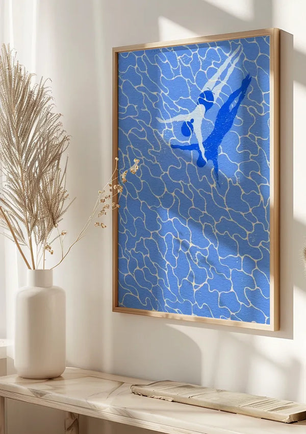 Een ingelijst schilderij De Blauwe Zwemmer van CollageDepot met een blauw abstract ontwerp met witte lijnen en een gestileerde figuur in beweging is op een witte muur gemonteerd. Naast het schilderij staat een witte vaas met gedroogde grassen, op een lichtgekleurde plank geplaatst.,Lichtbruin
