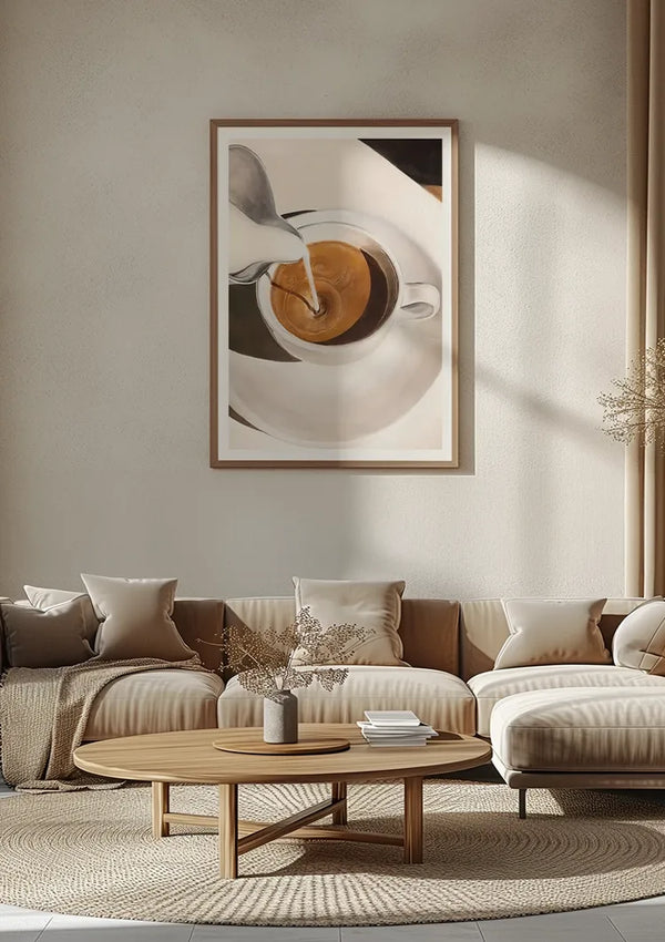 Een moderne woonkamer is voorzien van een groot Cappuccino-schilderij van CollageDepot van een koffiekopje waarin melk wordt gegoten, opgehangen boven een beige sofa met meerdere kussens. Voor de bank staat een ronde houten salontafel met daarop een vaas met gedroogde bloemen en een boek.,Lichtbruin