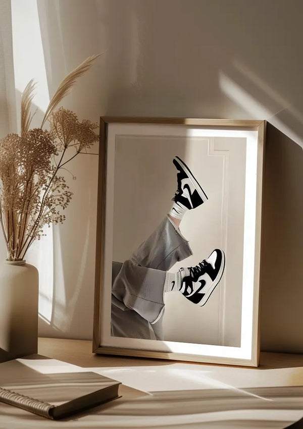 Ingelijste foto van benen met zwart-witte Nike Jordans, tegen een muur geleund, op een houten oppervlak geplaatst. Links staan gedroogde planten in een vaas, en natuurlijk licht filtert door vitrages, waardoor schaduwen werpen op de CollageDepot Nike Jordans Schilderij wanddecoratie.,Lichtbruin