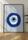 Een ingelijst abstract schilderij van een concentrisch cirkelpatroon rust tegen een witte muur op een lichte houten vloer. De cirkels, in de kleuren blauw, wit en zwart, lijken op een oog. Schaduwen van ruiten creëren kruisende lijnen over dit opvallende Turkse oogschilderij van CollageDepot.