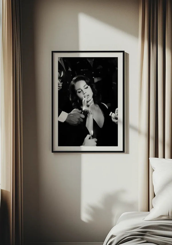 Een zwart-witte Glamorous Lana del Rey rokend van CollageDepot hangt aan een beige muur tussen twee lichtgekleurde gordijnen. De foto toont een persoon met gesloten ogen, die een sigaret vasthoudt terwijl een andere hand deze aansteekt. De rechteronderhoek van een netjes opgemaakt bed is zichtbaar.