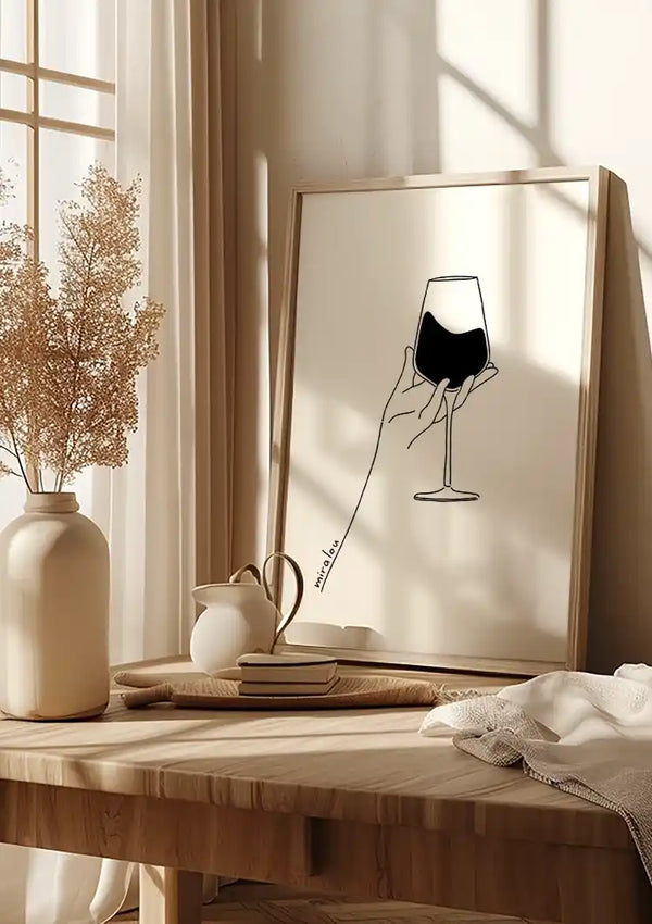 Een ingelijste lijntekening van een hand die een wijnglas vasthoudt, rust op een houten tafel versierd met **CollageDepot's Abstract Wine Glass Schilderij**. Op de tafel staat ook een vaasje met gedroogde bloemen, een theepot, twee boeken en een opgevouwen wit kleed. Zonlicht stroomt door een nabijgelegen raam en werpt schaduwen op deze elegante wanddecoratie-opstelling.,Lichtbruin