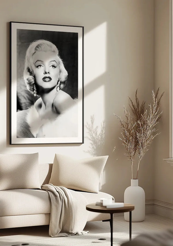 Een minimalistische woonkamer is voorzien van een beige bank met twee kussens, een lichte deken, een kleine ronde salontafel en een groot ingelijst Portret Marilyn Monroe Schilderij van CollageDepot. Een witte vaas met droog gras staat vlakbij als smaakvolle wanddecoratie.,Zwart