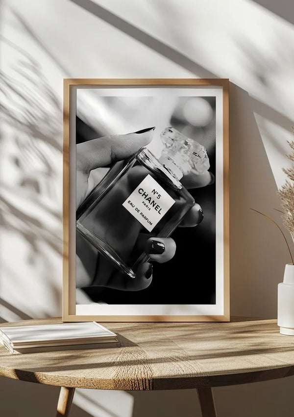 Op een houten tafel staat een ingelijste zwart-witfoto van een hand die een Parfum In Hand Schilderij-fles van CollageDepot vasthoudt. Op de tafel staat ook een witte vaas met gedroogd blad en een stapel boeken, met schaduwen van een nabijgelegen plant op de muur, waardoor een elegante wanddecoratie ontstaat.,Lichtbruin