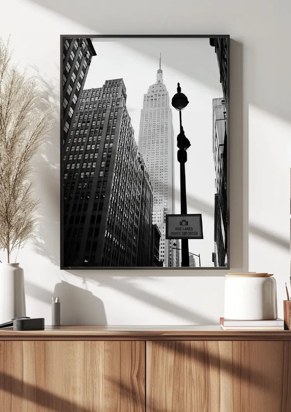 Een zwart-wit Streets of New York-schilderij van CollageDepot met wolkenkrabbers, waaronder het Empire State Building, hangt aan een witte muur boven een lichte houten kast. Op de kast staat een witte vaas met gedroogd pampagras en andere kleine decoratieve spulletjes, beveiligd door een magnetisch ophangsysteem.