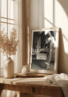 Een houten tafel bij een raam is versierd met een ingelijste Givenchy-poster, vazen met delicate droge planten en een kleine theepot bij een stapel boeken. Het zonlicht stroomt door transparante gordijnen en werpt zachte schaduwen over de tafel en wanddecoratie met een elegant Givenchy-schilderij van CollageDepot.,Lichtbruin