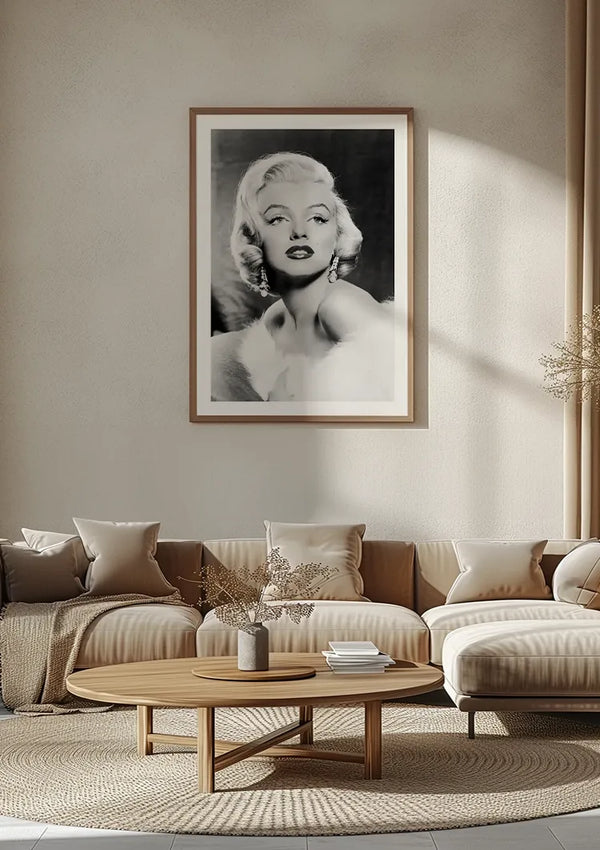 Een zwart-wit portret Marilyn Monroe schilderij van Marilyn Monroe met kort blond haar en elegante kleding is ingelijst en opgehangen aan een beige muur. Onder de foto ziet u een moderne woonkameropstelling met een beige bank, ronde houten salontafel en diverse kussens. Het kunstwerk is van CollageDepot.,Lichtbruin