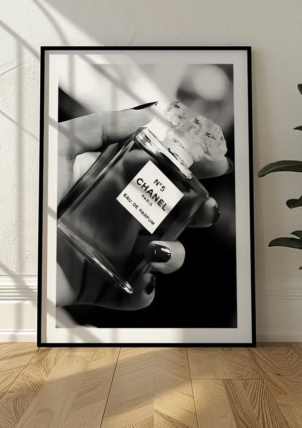 Zwart-witfoto van een hand die een Parfum In Hand Schilderij CollageDepot-fles vasthoudt, ingelijst als een prachtig zwart-wit schilderij en rustend op een houten vloer tegen een witte muur. De hand is voorzien van donkere nagellak en de afbeelding wordt prominent weergegeven door middel van een magnetisch ophangsysteem.,Zwart