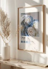 Een ingelijste CollageDepot Lichtblauwe En Witte Rozen Cloé Parijs Schilderij, een prachtige wanddecoratie, hangt aan een witte muur. De poster toont delicate lichtblauwe en witte rozen met "Dior" en "Paris, Frankrijk" onder de afbeelding. Daaronder staat een strakke witte vaas met gedroogde bloemstukken op het oppervlak.,Lichtbruin